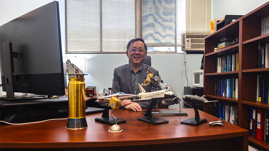 Ping Lu, SDSU professor of aerospace engineering seated at desk with spacecraft models, computer monitor, and bookshelf in an office setting.
